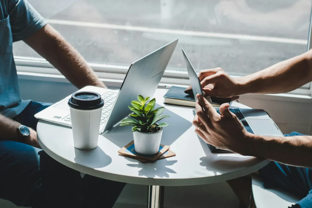 Two individuals seated at a table, each using a laptop, engaged in a collaborative work session.