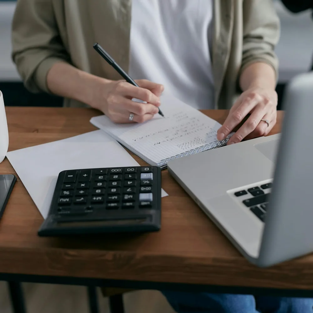 A woman diligently writes in a notebook with a pen while seated at her desk, immersed in her work.